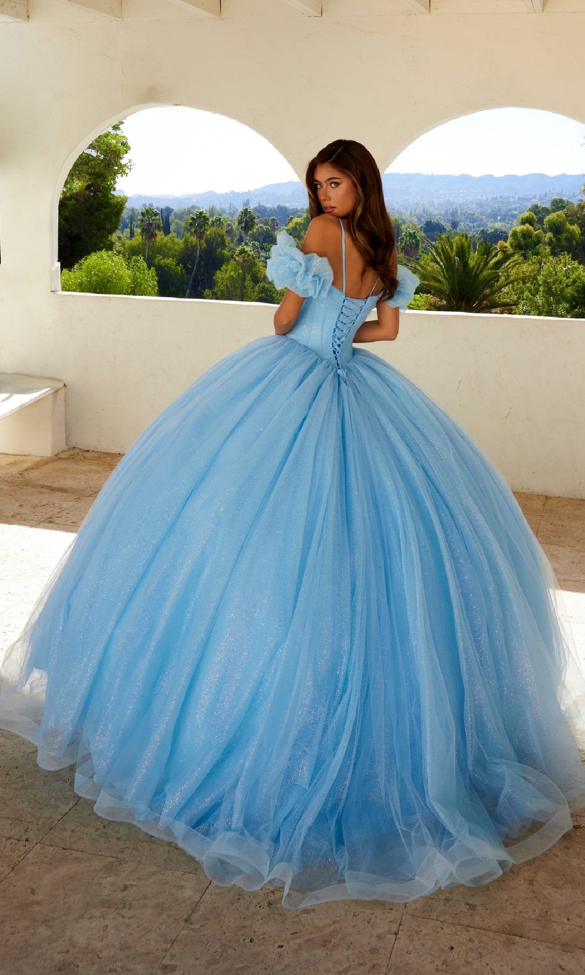 A young woman stands on a terrace in the Quinceanera Dress JT1487H By Juliet, a glitter tulle ball gown with puff off-the-shoulder sleeves, gazing over her shoulder at the trees and hills beyond.